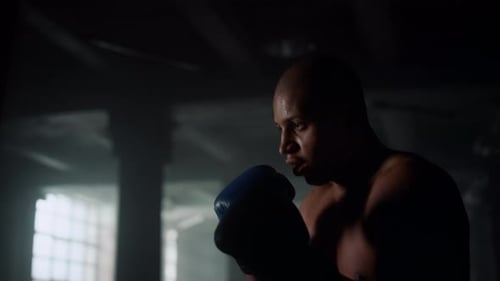 African american boxer training kicks on punch bag in dark loft gym
