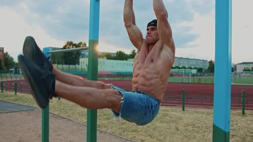 Young Muscular Man Without Tshirt is Training at City Stadium and Doing Hanging Leg Swings on