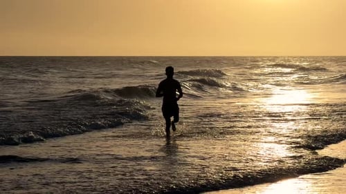 Young man running along the beach towards the camera during sunset.