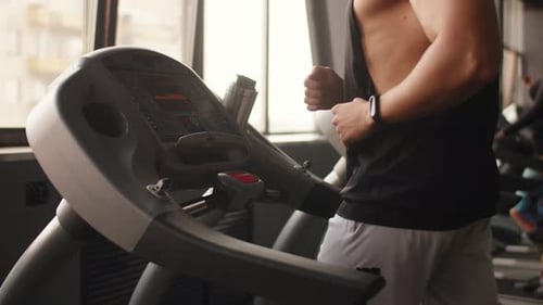 Side View of a Muscular Man in a Tank Top and a Fitness Bracelet Runs on a Treadmill in a Fitness