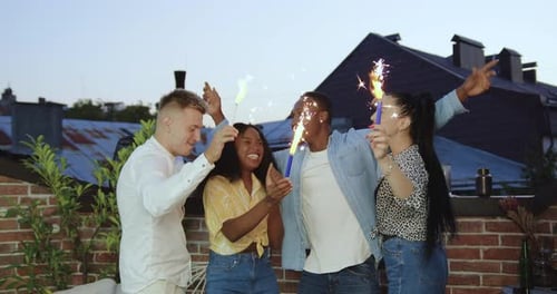 Happy young male and female friends celebrating summer party on terrace with sparkled lights at sund