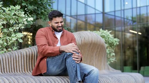 Businessman suffers from painful severe knee joint pain sitting on a bench on the street near office