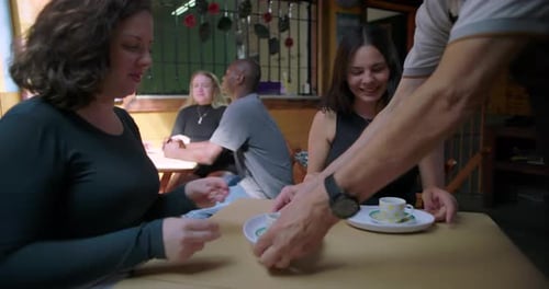 Friends receiving coffee at cafe table as server places cups by hand, casual hospitality moment,