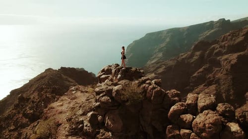 A woman stands on a rocky outcrop overlooking the ocean and cliffs during a sunny day.