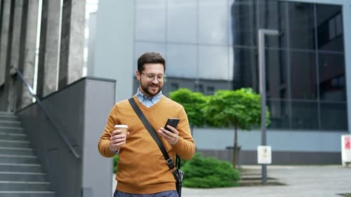 Smiling businessman walking outdoors using phone, holding coffee, while browsing or chatting online.