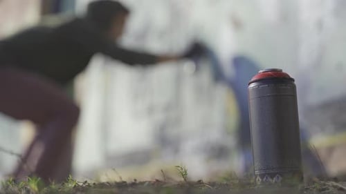 Homem desfocado pintando grafite na parede ao fundo, em primeiro plano, frasco de spray em pé