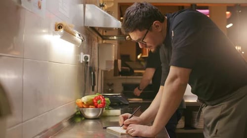 Professional Chef Writing Recipe Notes at Workstation in Restaurant Kitchen