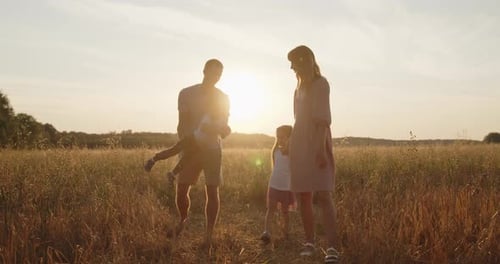 Young Smiling Family of Four Walk Through a Golden Field at Sunset Together