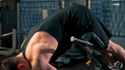 in the Gym Young Handsome Guy with Tattoos in a Black Tshirt Shakes His Back on an Exercise Machine