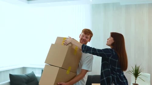 Young Caucasian Redhaired Guy Holding Many Cardboard Boxes in Hands and Woman Putting Another One on