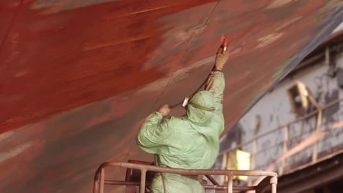 Worker in Protective Gear Applies Red Paint to Ship Hull in Dry Dock Industrial Painting Maintenance