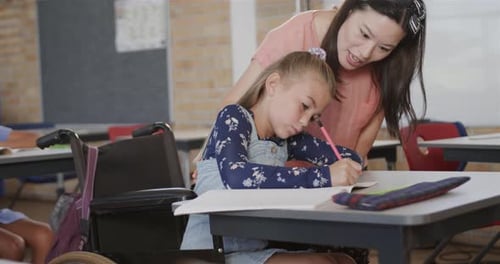 Diverse female teacher teaching schoolgirl in wheelchair writing in classroom at elementary school