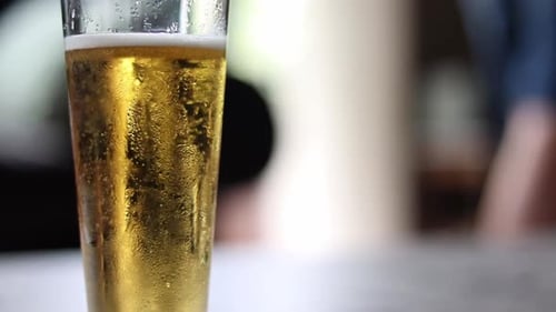 a Golden beverage bubbling in a glass with foam, while people walk by in the background.