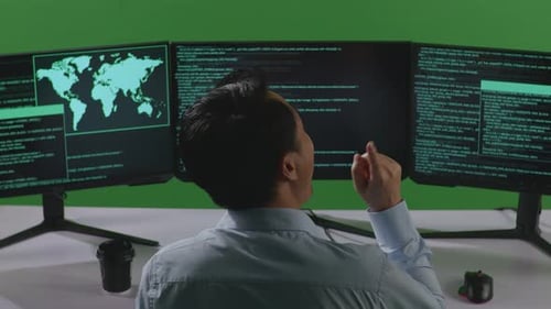 Back View Of Asian Man Thinking While Write Code With Multiple Computer Screens In Green Screen