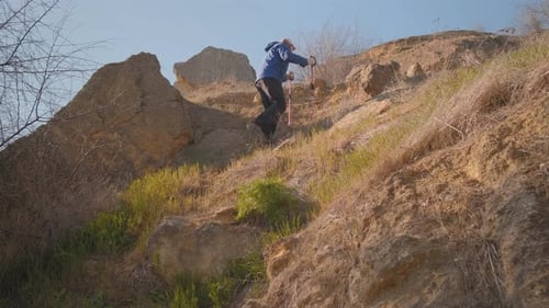A Tourist Hiker Climbs A Rocky Slope With Hiking Sticks
