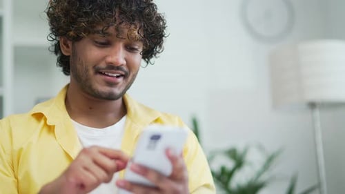 Happy handsome young man in yellow shirt uses phone in home office room Mixed race male typing brows