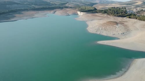 Luftaufnahme über dem türkisfarbenen Wasser und Sand eines Stausees mit einem Damm im Hintergrund.