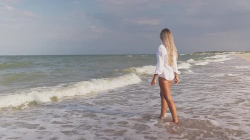 A Slender Girl in a Gentle Blue Swimsuit and Shirt Walks Along the Sandy Beach Near the Blue Sea