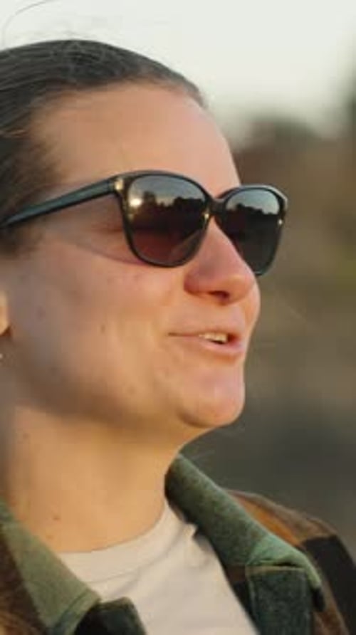 Une jeune femme portant des lunettes et une chemise à carreaux dans la forêt regarde le coucher de soleil en riant Vertica