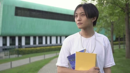 Asian Student with Textbooks Outdoors Against the Backdrop of a College or University