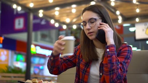 A Young Woman in a Cafe is Talking on the Phone and Drinking a Hot Drink
