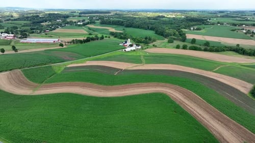 Aerial view of curving agricultural fields with farm buildings, surrounded by greenery. Summer in Am