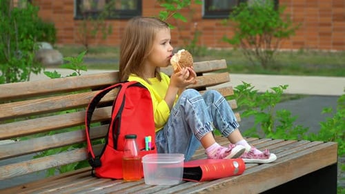 Student Girl Sits with Backpack and Eats Hamburger Schoolchild Has Breakfast