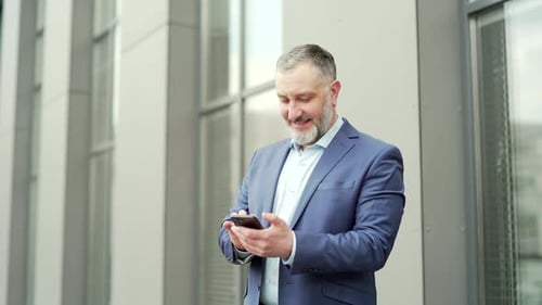 Happy mature business man in formal suit using smartphone while standing outside near the office