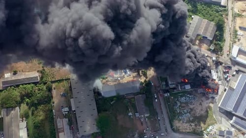 Dramatic aerial view of a factory engulfed in flames, with thick black smoke rising as fierce flames