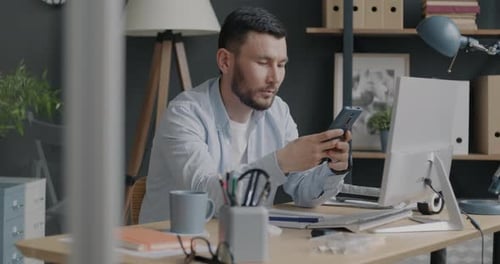 Portrait of Biracial Entrepreneur Using Smart Phone Messaging Sitting at Computer Desk in Office