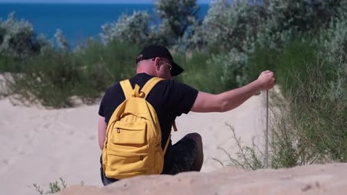 Man with Backpack Sits on Sand Beach Dune Sand Puffs Through His Fingers