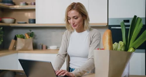Smiling Woman Using Laptop with Groceries in Kitchen