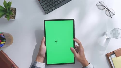 Top down view of hands holding a mock up green screen tablet on a white desk