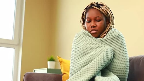 Mujer afro envuelta en una manta azul y bebiendo té caliente en la sala de estar