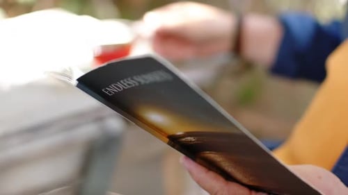 Right hand is initiating fanning ENDLESS SUNSETS booklet pages to reveal title on porch near cup