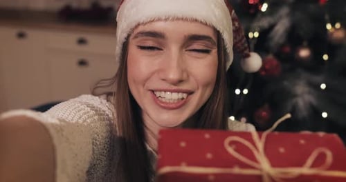 Smiling Woman Wearing Santas Hat Sitting Near Christmas Tree at Home Holding and Shaking Red Gift