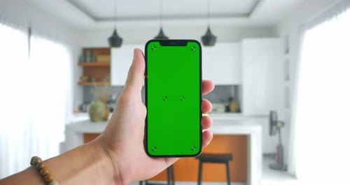 Male Hand Holding Smartphone with Green Screen Indoors on Modern Kitchen Background