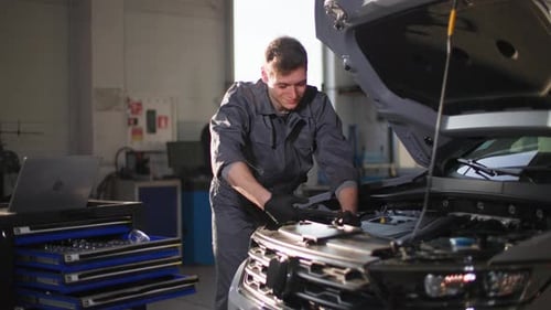 Young Man in Uniform with Tool in His Hands Checks Car Engine Near Open Hood at Station Smiling and