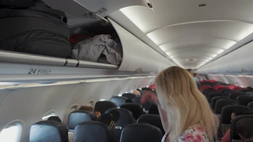 Une femme blonde tend la main pour ouvrir le compartiment supérieur et fourre un grand sac à dos pour le ranger