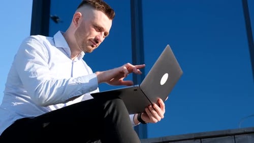 A Young Businessman Works on a Laptop Outdoors Against the Background of a Corporate Glass Office