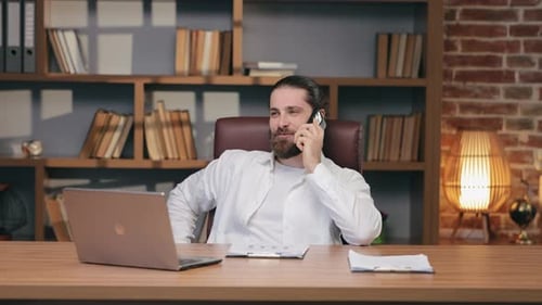 Confident Caucasian Man with Wellgroomed Beard Talking on Smartphone While Sitting at Desk in Office
