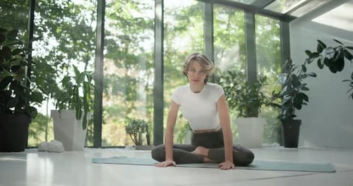 Young Caucasian Fitness Woman Practicing Yoga on a Yoga Mat in a Modern Studio with Plants and Large