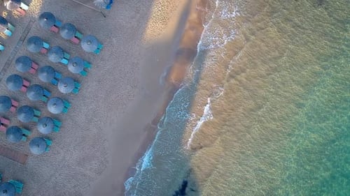 Aerial Top Down View Over Waves Crashing On Beach With Empty Sunbeds With Blue Parasols