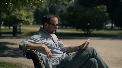 Smiling man enjoys movie on tablet computer in sunny city park bench relaxation