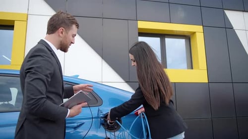 Businessman in a dealership shows a girl how to charge an electric car at a charging station