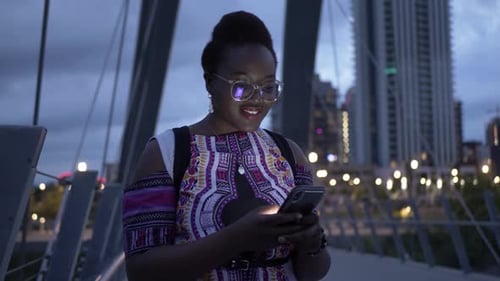 Stylish young woman uses smart phone on urban bridge at night