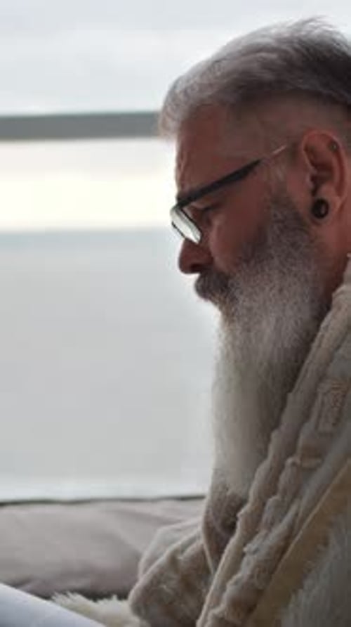 A Focused Man with Gray Hair and a Beard Wearing Glasses Reads a Magazine Wrapped in a Blanket on