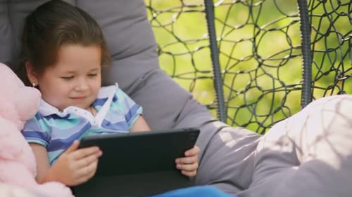 Girl Sits in a Rocking Chair Cocoon with Looks at the Tablet