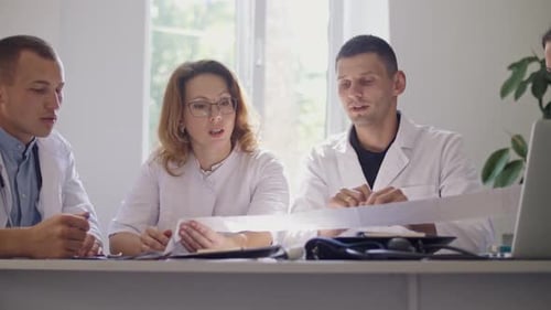Team of Four Doctors in White Coats Sitting at Table in Hospital and Working With a Laptop