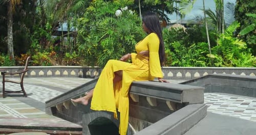 A girl in a buttery yellow dress sitting on the bridge at the grand entrance of a timeless castle.